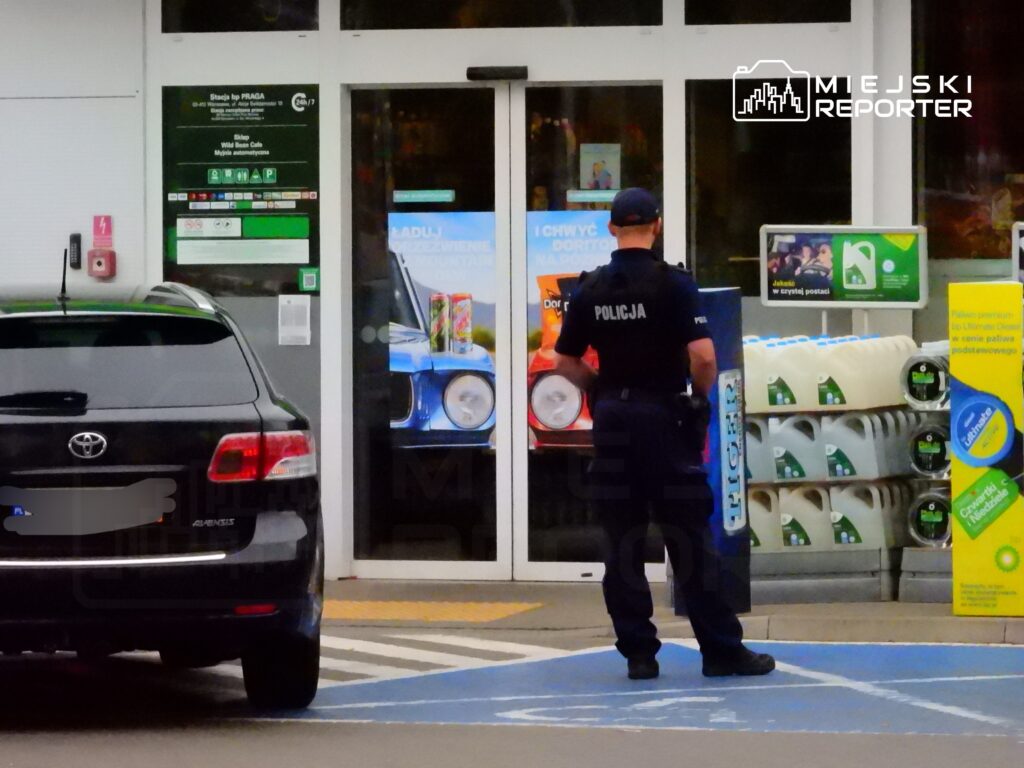 Policjant w mundurze stoi przed wejściem do stacji benzynowej, obok samochodu osobowego.