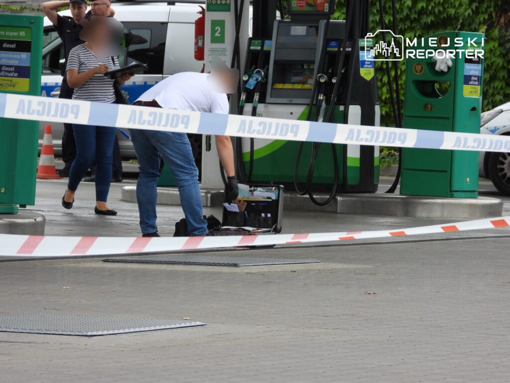 Mężczyzna w rękawiczkach bada teren przy stacji paliw, podczas gdy kobieta z notatnikiem obserwuje sytuację.