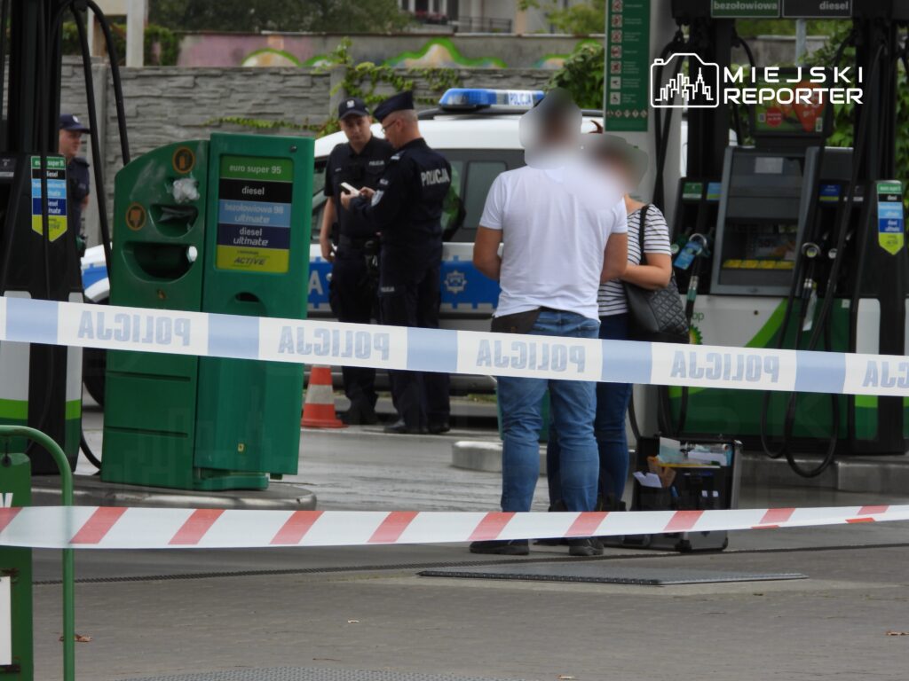 Dwóch policjantów rozmawia z dwiema osobami przy stacji benzynowej, otoczeni taśmą policyjną.
