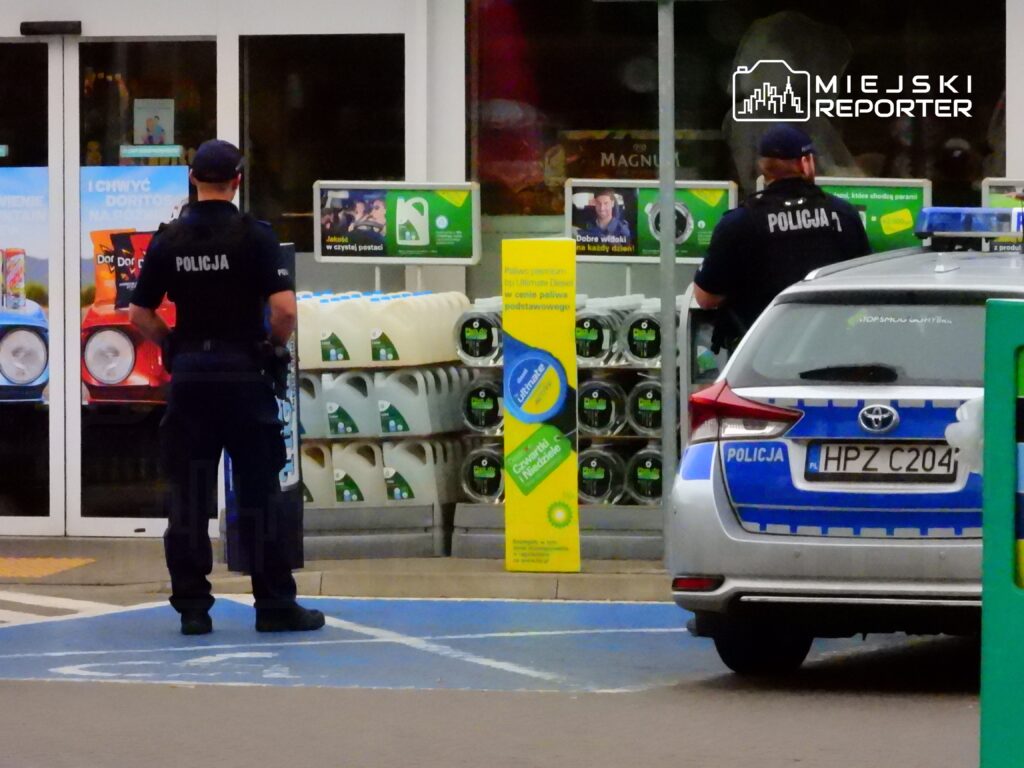 Dwóch policjantów w mundurach stoi przed wejściem do sklepu, obok radiowozu policyjnego.