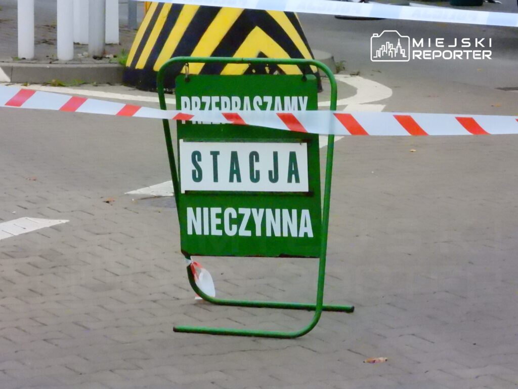 Zielony znak ostrzegawczy z napisem "STACJA NIECZYNNA" otoczony taśmą zabezpieczającą, ustawiony na ulicy.