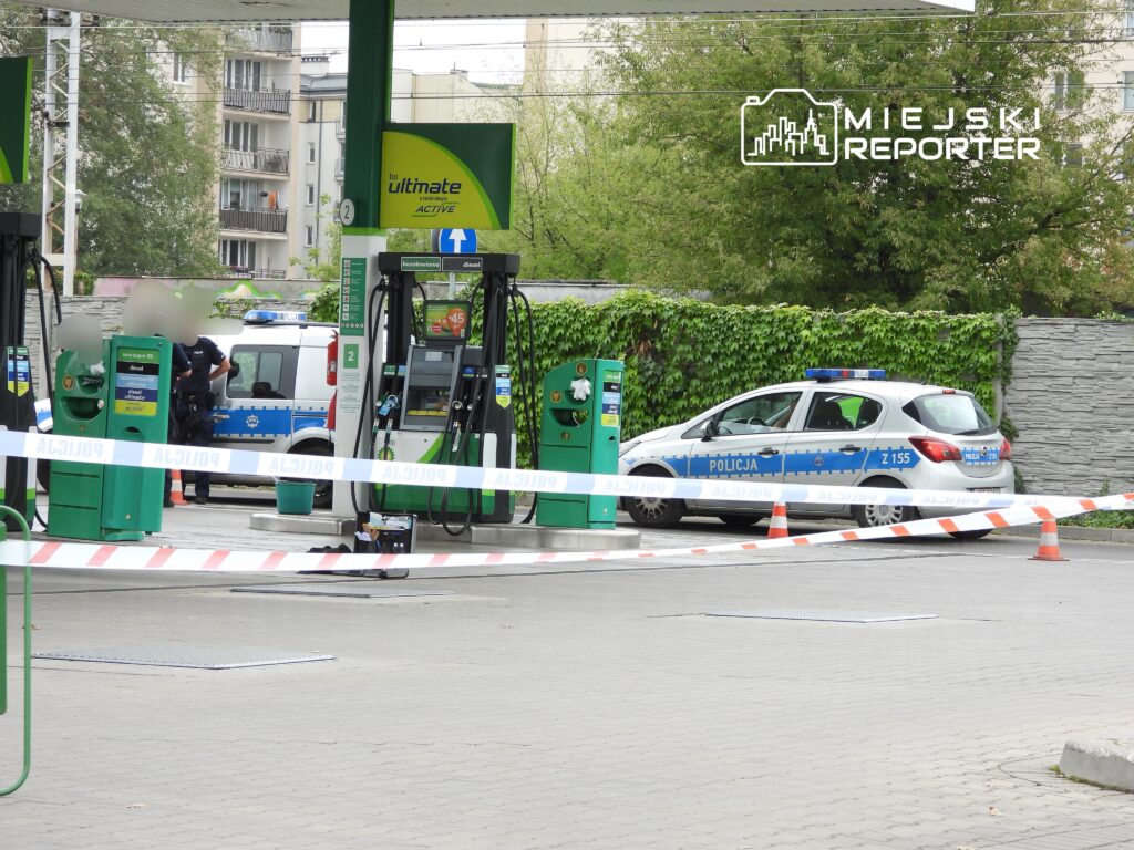 Policjant rozmawia z kolegą przy stacji paliw, w tle oznakowany radiowóz oraz taśmy zabezpieczające.