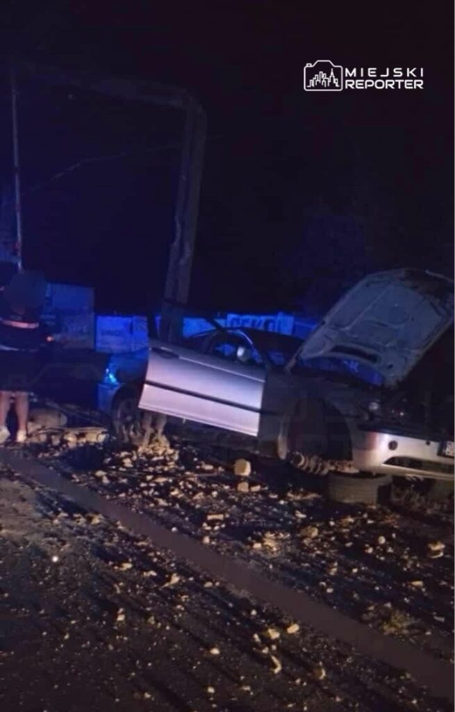 Mężczyzna w kamizelce odblaskowej bada uszkodzony samochód na drodze w nocy, obok leżą odłamki.