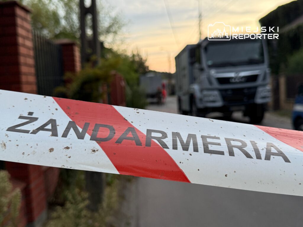 Czerwono-biała taśma z napisem "ŻANDARMERIA" w tle, na drodze przejeżdża ciężarówka.