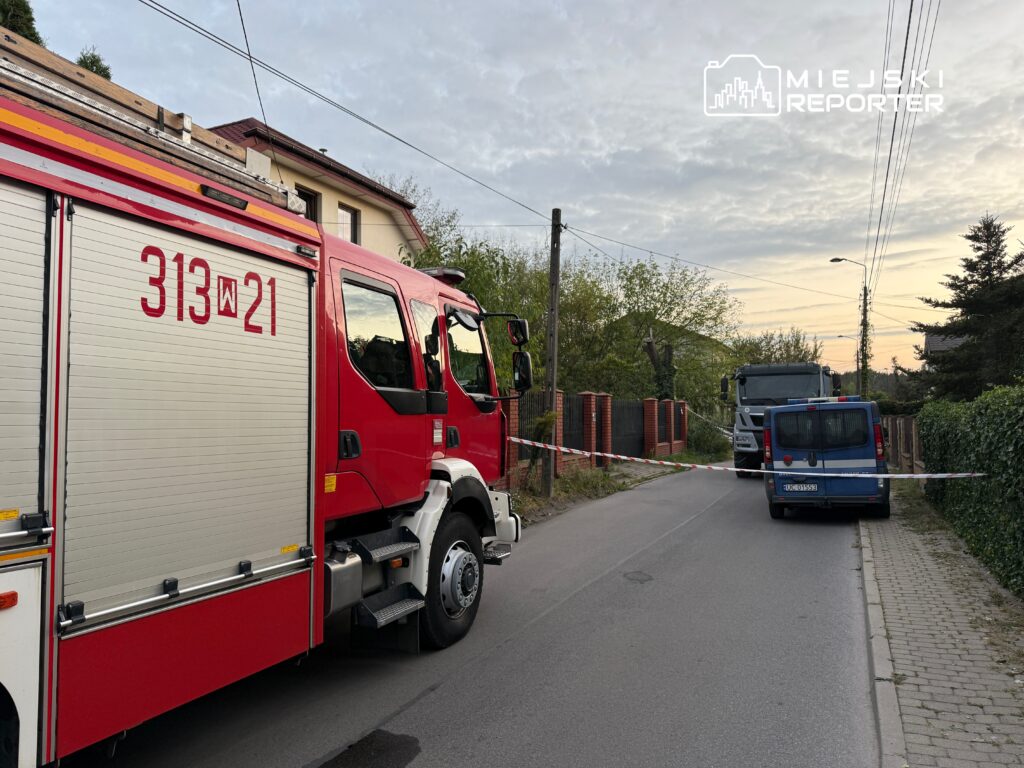 Czerwony wóz strażacki stoi na drodze, obok niebieski furgon, a teren jest zabezpieczony taśmą ostrzegawczą.