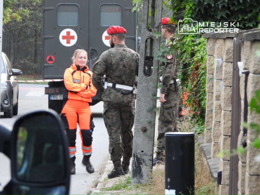 Kobieta w pomarańczowym uniformie rozmawia z żołnierzem w mundurze na ulicy obok wojskowego pojazdu.