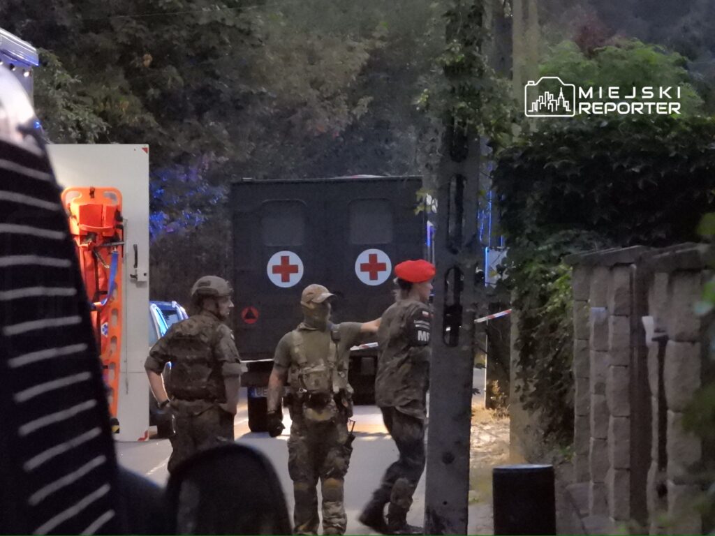 Trzech żołnierzy w mundurach omawia sytuację przy wojskowym pojeździe z czerwonym krzyżem w tle.