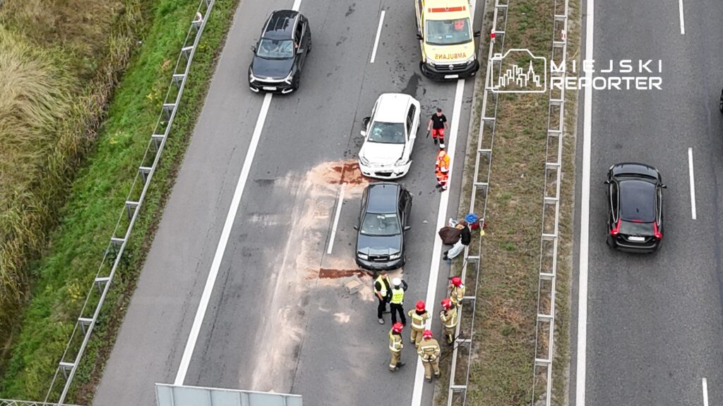 Na drodze doszło do kolizji dwóch samochodów; obok pracują strażacy i ratownicy medyczni.