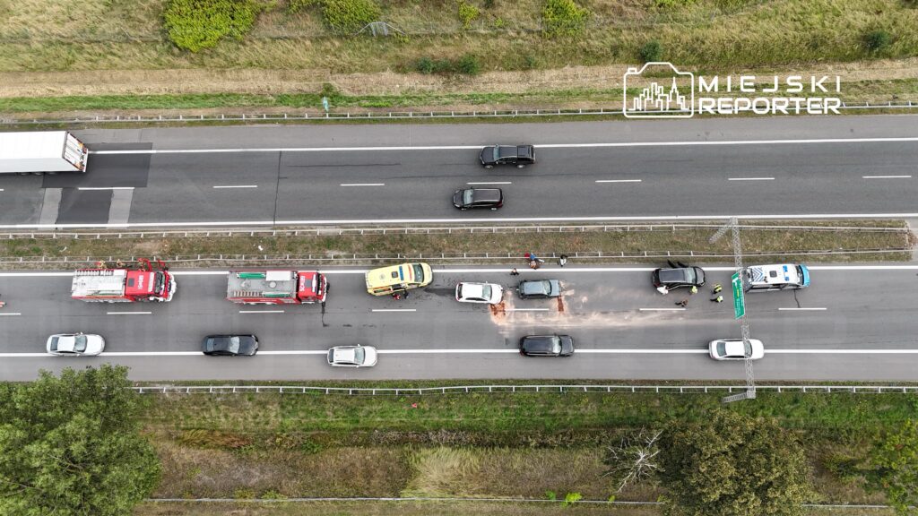 Na drodze znajdują się trzy samochody, w tym pojazd z uszkodzeniem, obok stoją ratownicy i policjanci.