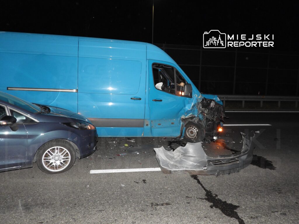 Niebieski furgon z uszkodzoną przednią częścią stoi na drodze, obok niego leży zniszczony fragment zderzaka.