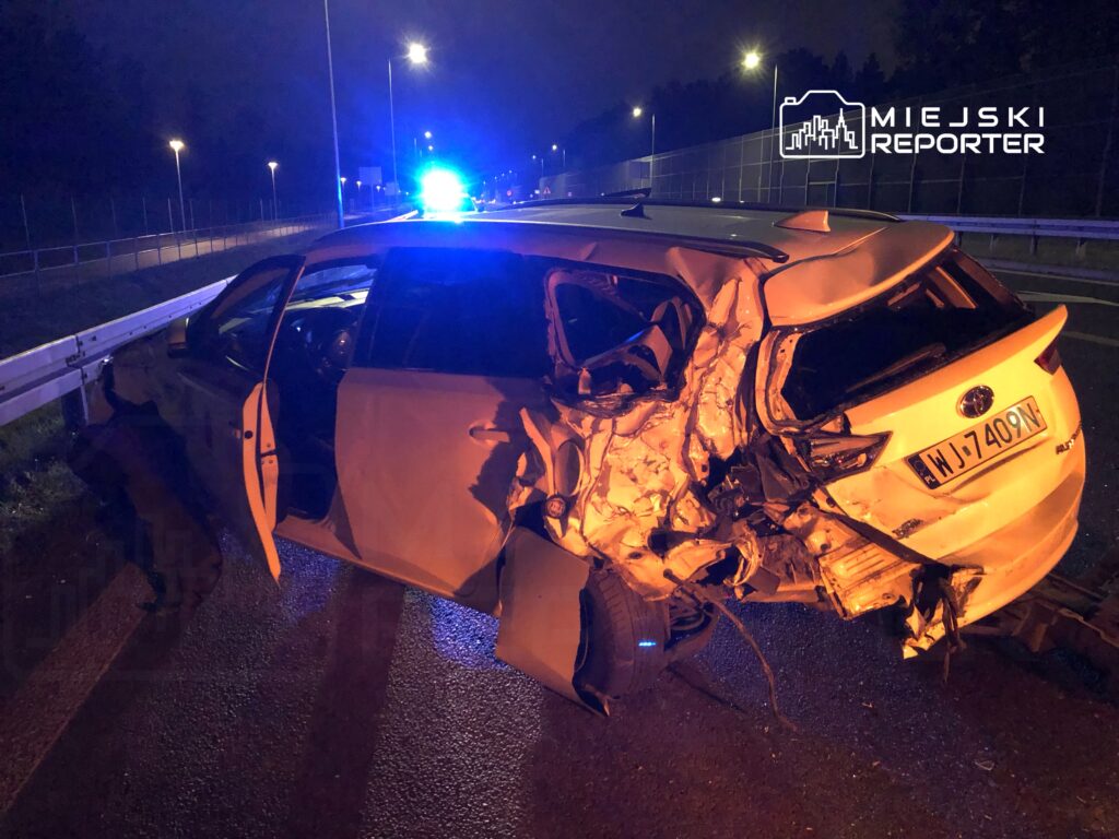 Uszkodzony samochód osobowy stoi przy barierce na drodze, oświetlony niebieskimi światłami policyjnymi w nocy.