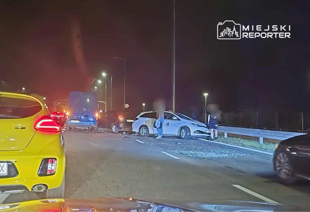Zdarzenie drogowe z udziałem kilku pojazdów na oświetlonej ulicy w nocy, z osobami sprawdzającymi uszkodzenia.
