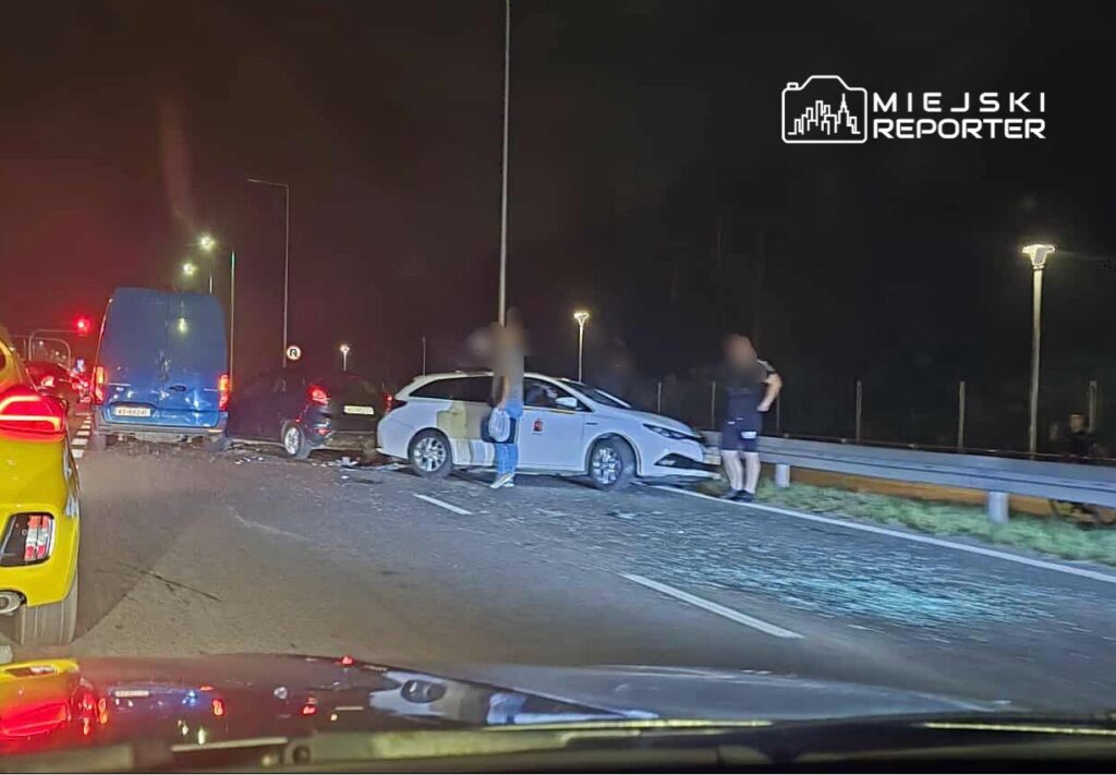 Kolizja drogowa z udziałem kilku pojazdów na nocnej jezdni, z uszkodzonymi samochodami i stojącymi obok osobami.