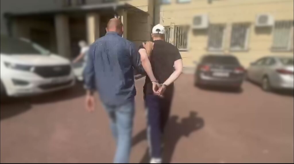 Mężczyzna w kajdankach prowadzony przez funkcjonariusza na parkingu przed budynkiem.