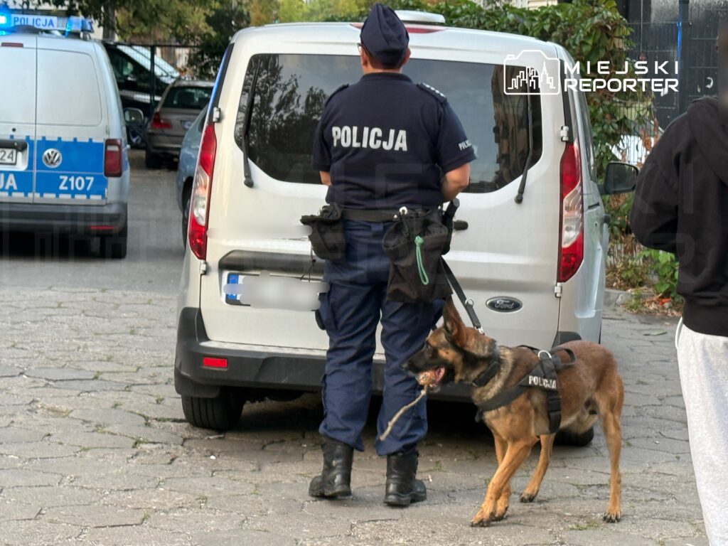 Policjant z psem służbowym stoi obok białego pojazdu, w tle widoczne radiowozy policyjne.