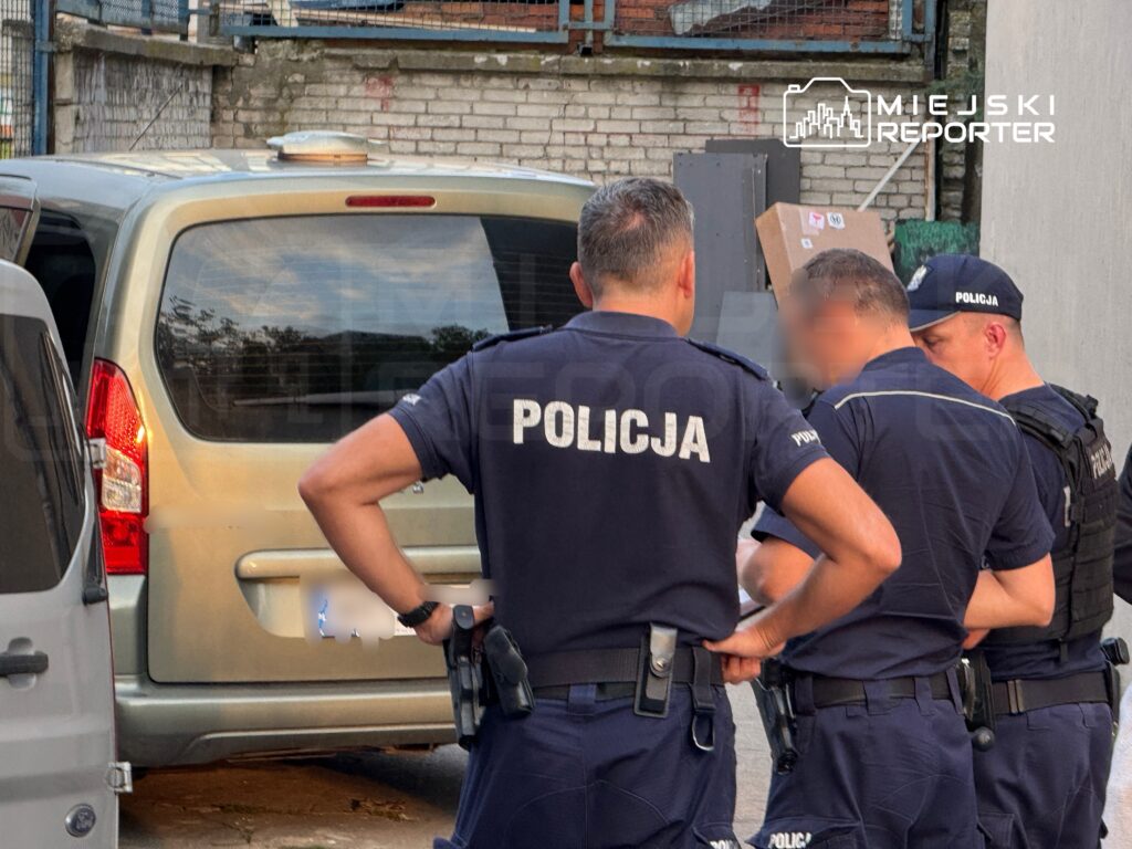 Grupa policjantów omawia sytuację przy zaparkowanym samochodzie w miejskim otoczeniu.