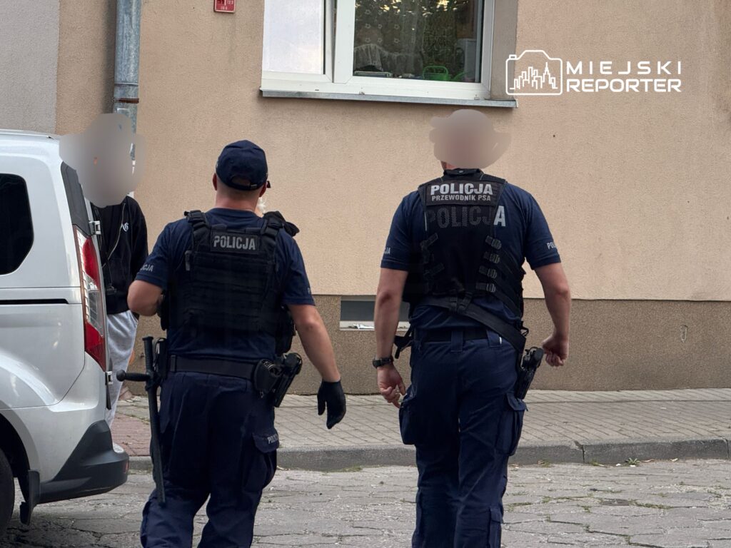 Dwóch policjantów w mundurach, jeden z psem, idzie w kierunku białego furgonu na ulicy.