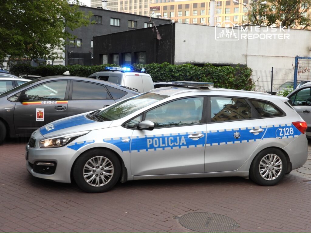 Policyjny radiowóz zaparkowany na ulicy, obok innych samochodów, z włączonymi sygnałami świetlnymi.
