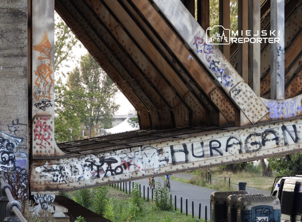 Metalowa konstrukcja mostu z graffiti na przęsłach, w tle ścieżka rowerowa i zieleń.