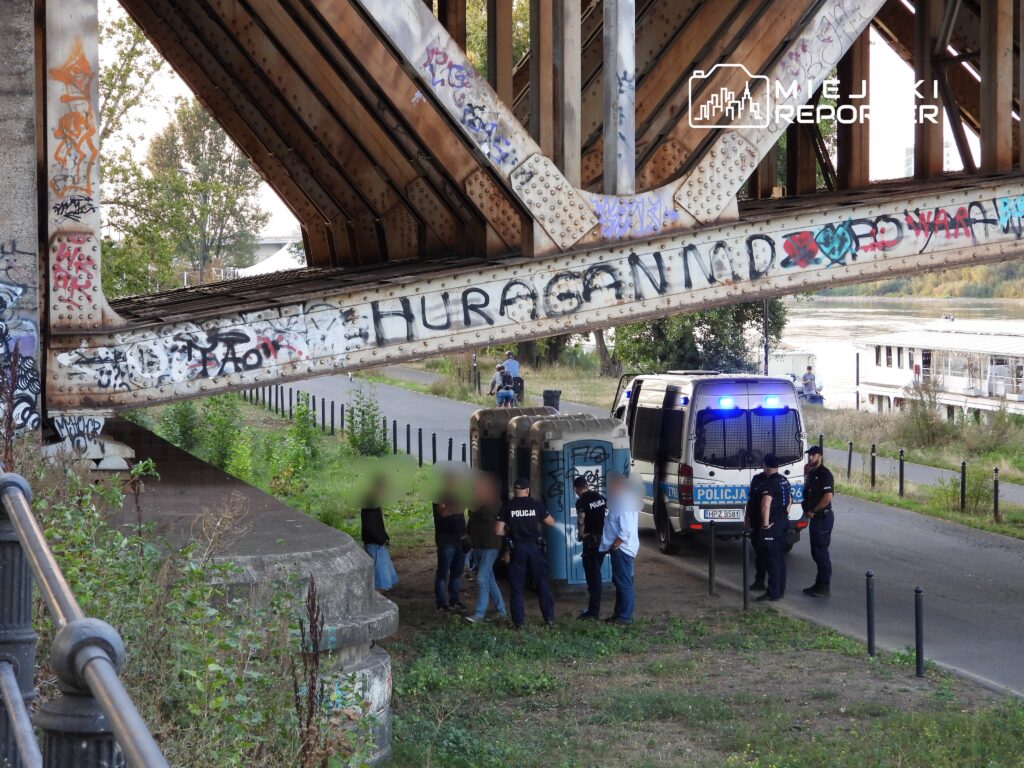 Grupa policjantów rozmawia z kilkoma osobami pod mostem, obok stoi policyjny radiowóz z włączonymi światłami.