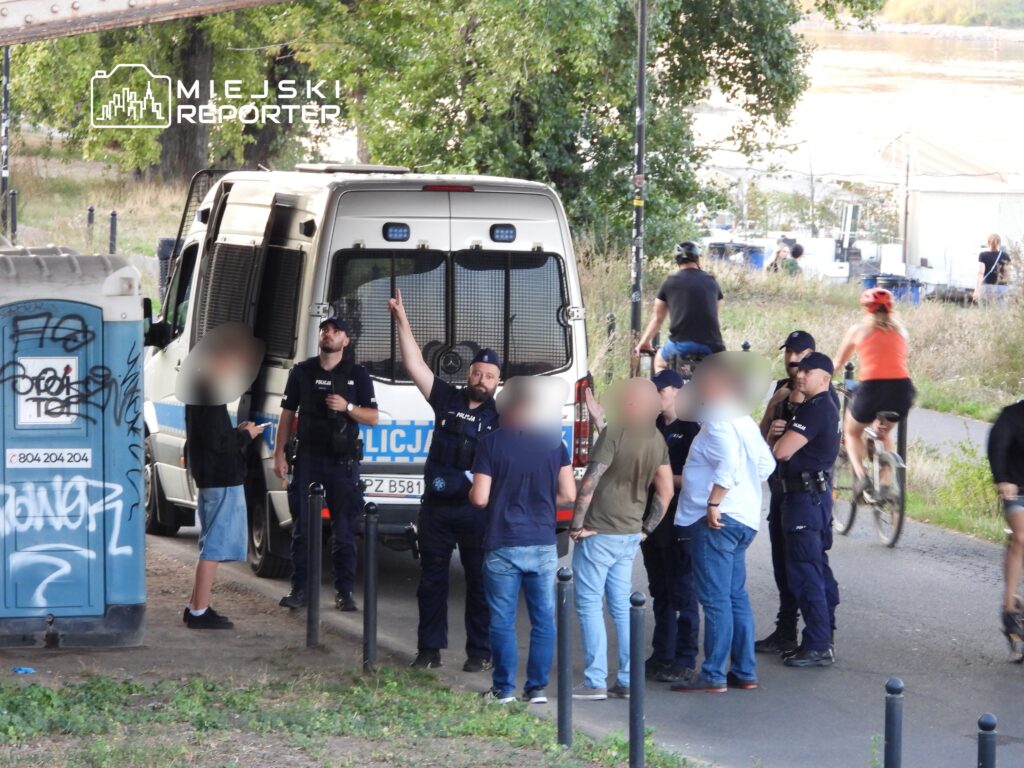 Grupa policjantów w mundurach rozmawia z mężczyzną na ścieżce rowerowej obok radiowozu. W tle przejeżdżają rowerzyści.