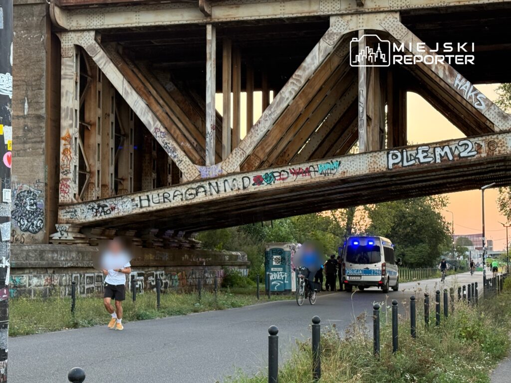 Mężczyzna w białej koszulce biega pod graffiti pokrytym mostem, obok zatrzymany radiowóz policyjny oraz rowerzyści.