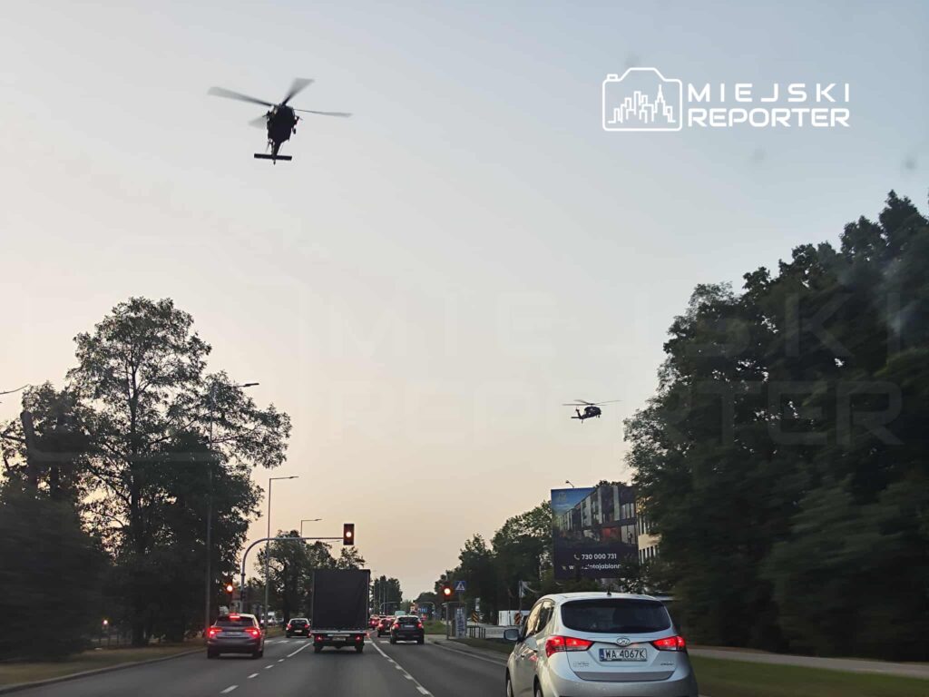 Dwa helikoptery przelatują nad ruchliwą ulicą, obok pojazdów w korku i sygnalizacji świetlnej.
