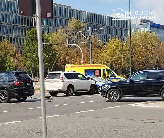 W ruchu drogowym doszło do kolizji dwóch samochodów osobowych, obok stoi karetka pogotowia. W tle nowoczesny biurowiec.