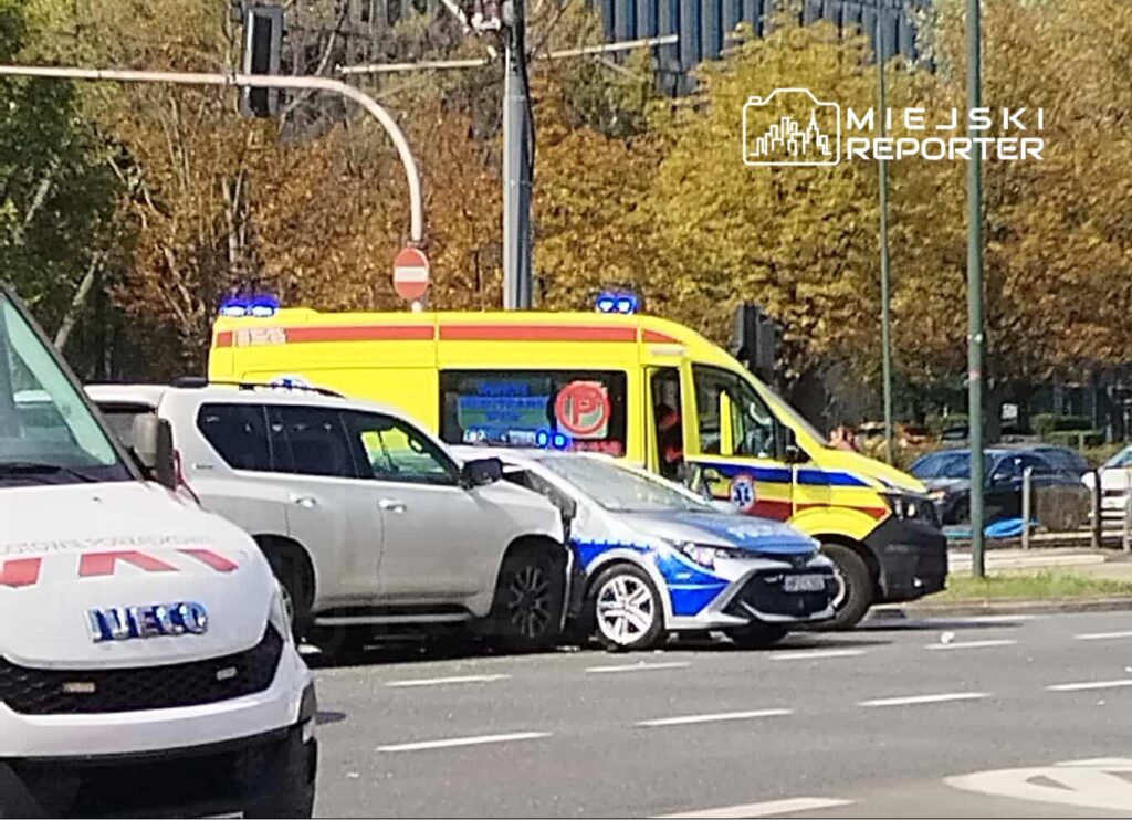 Na skrzyżowaniu stoi ambulans z włączonymi sygnałami świetlnymi, obok uszkodzone samochody osobowe.