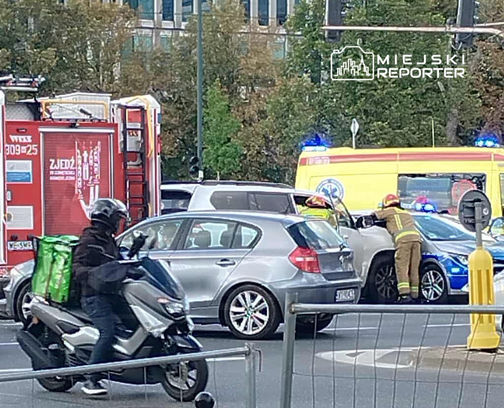Kierowca w mundurze strażackim sprawdza uszkodzone auto na skrzyżowaniu, obok stoi karetka i inne pojazdy.