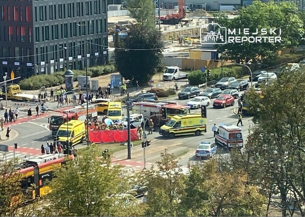 Wielka akcja służb ratunkowych na skrzyżowaniu, z wieloma ambulansami i pojazdami straży pożarnej, w otoczeniu ruchu miejskiego.