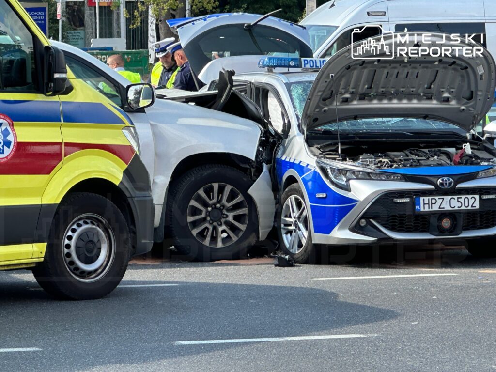 Wypadek drogowy z udziałem białego SUV-a i nieoznakowanego radiowozu, obok stoi karetka pogotowia oraz policjanci.