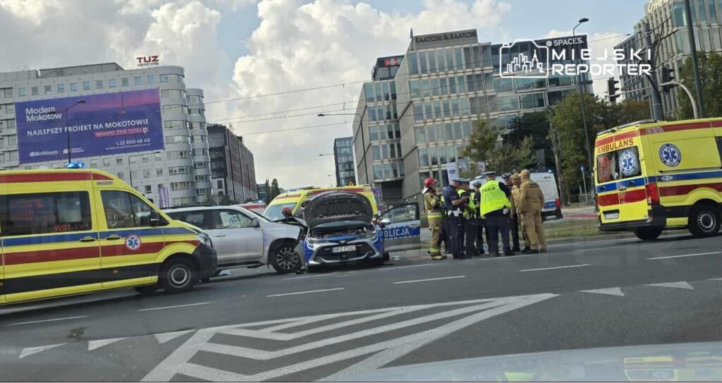Wielu ratowników i policjantów zbiera się wokół zderzonych pojazdów na ruchliwej ulicy.