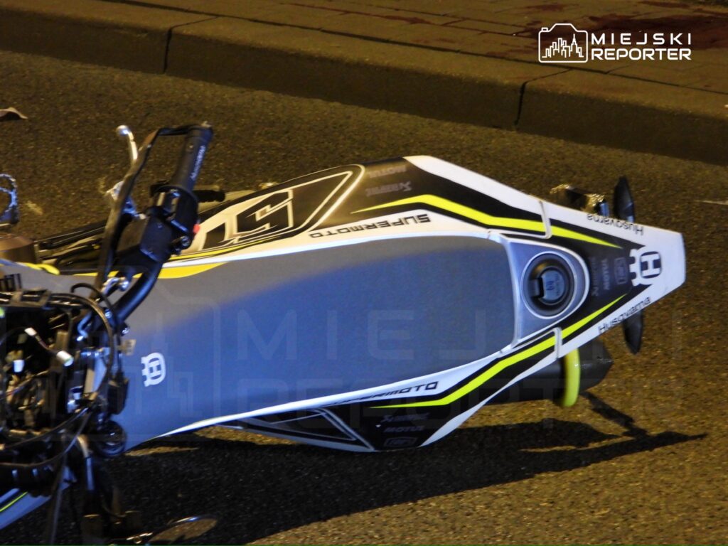 Motocykl leżący na boku na drodze, w nocnym otoczeniu, z widocznymi uszkodzeniami.