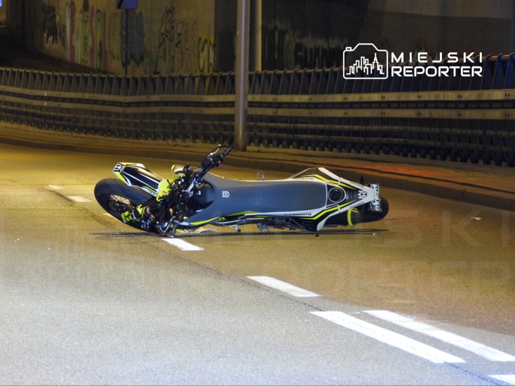 Motocykl leżący na boku na oświetlonej jezdni, otoczony graffiti na murze w tle.