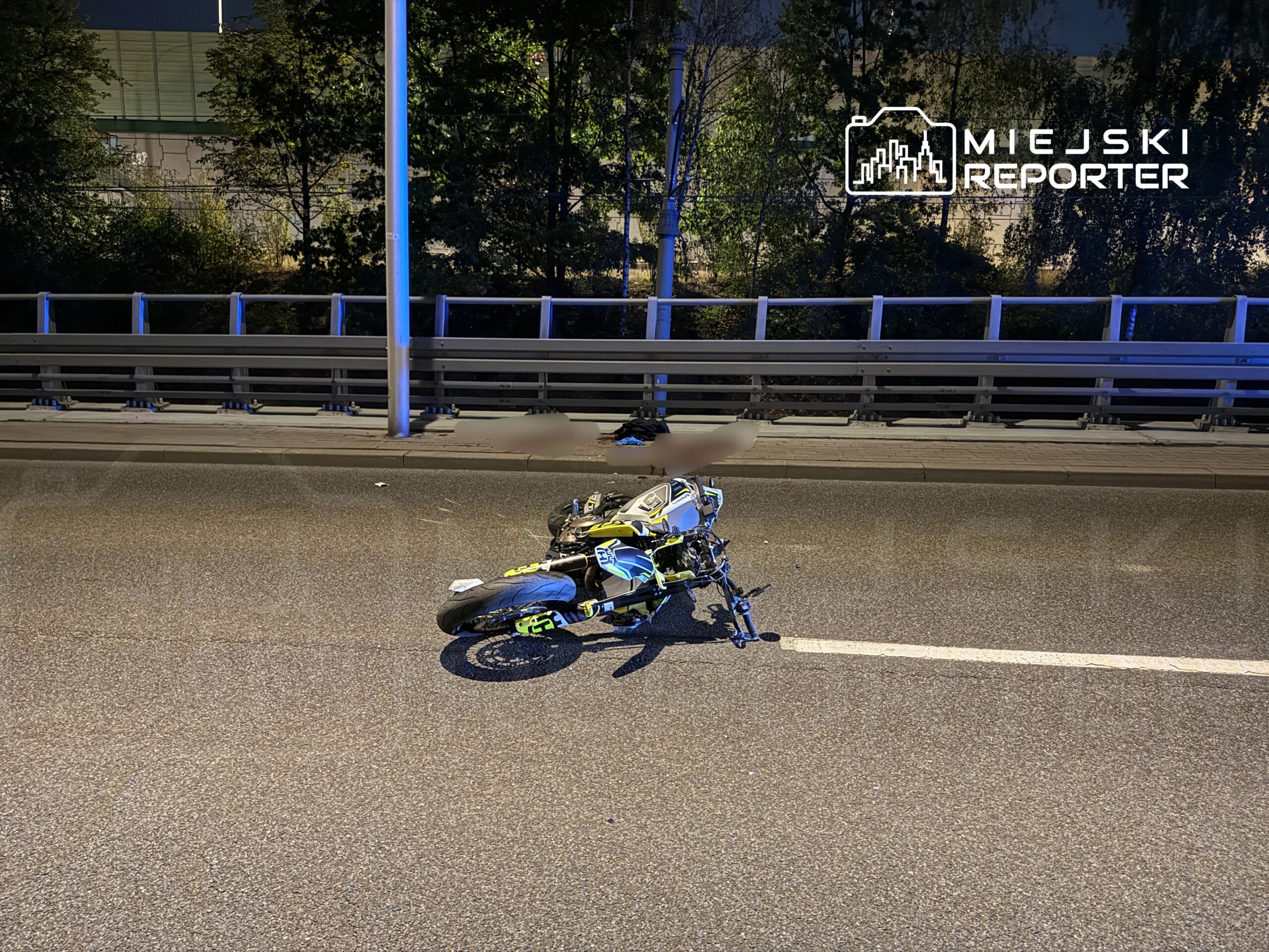Motocykl leżący na jezdni, obok bariery ochronnej, w otoczeniu drzew i oświetlenia ulicznego.