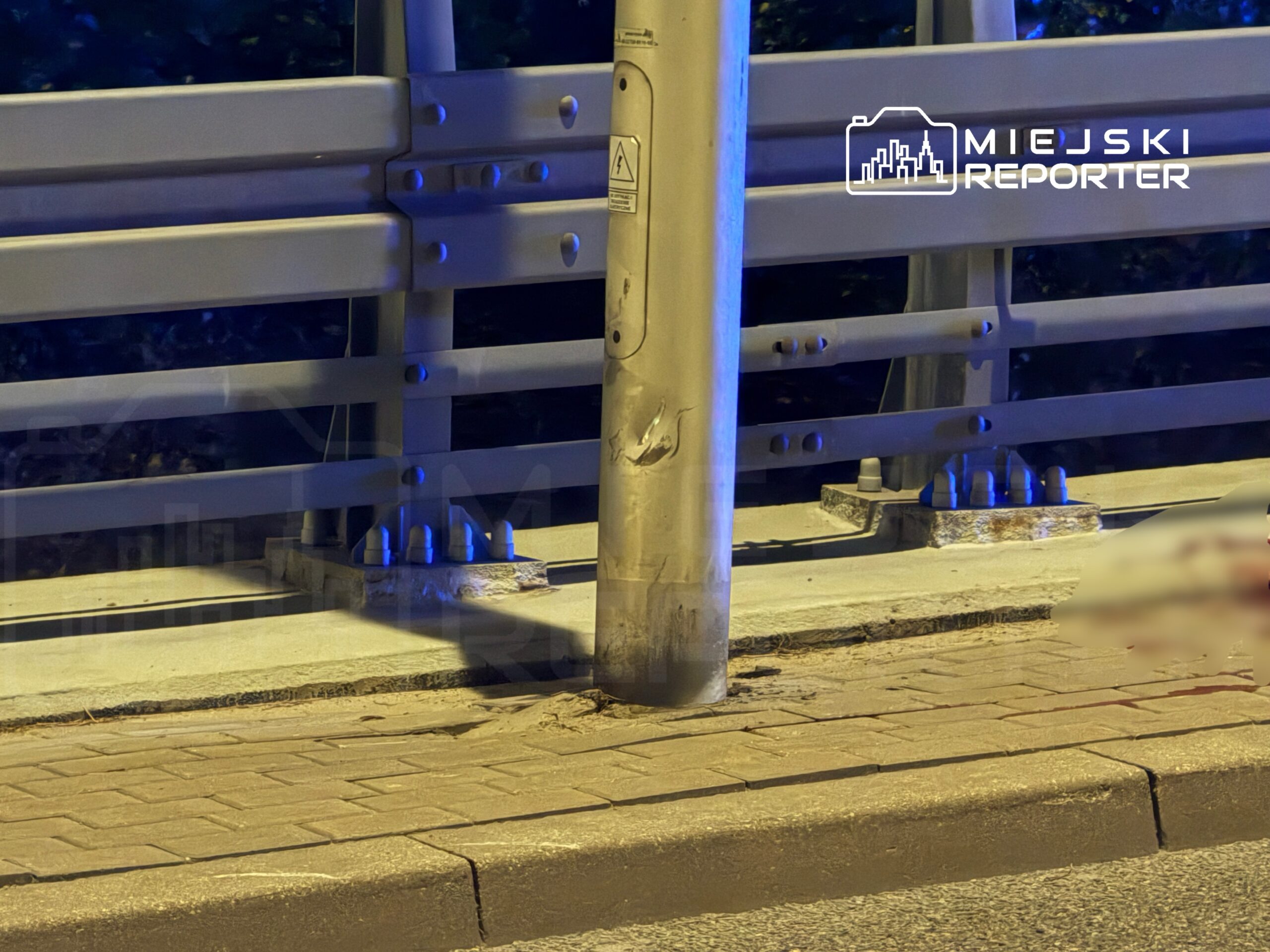 Metalowy słup oświetleniowy z uszkodzeniami, stojący na chodniku obok stalowej balustrady.