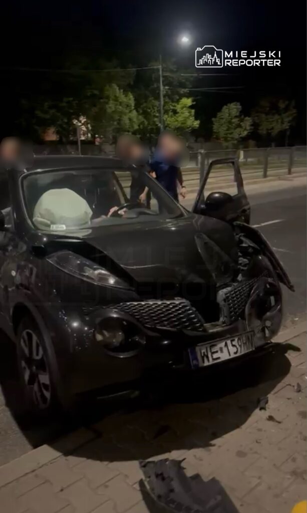 Czarny samochód osobowy z uszkodzoną przednią częścią stoi na chodniku, obok dwóch osób rozmawiających.