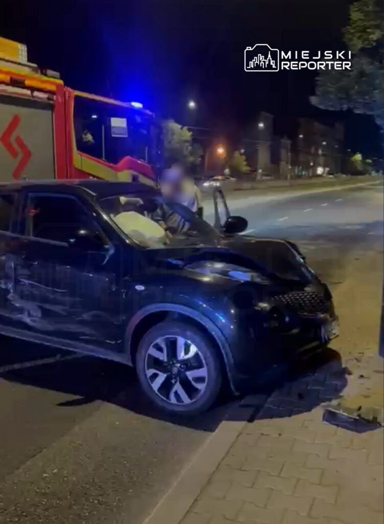 Czarny samochód osobowy z uszkodzoną przednią częścią stoi przy drodze, obok wozu strażackiego z włączonymi sygnalizacjami.