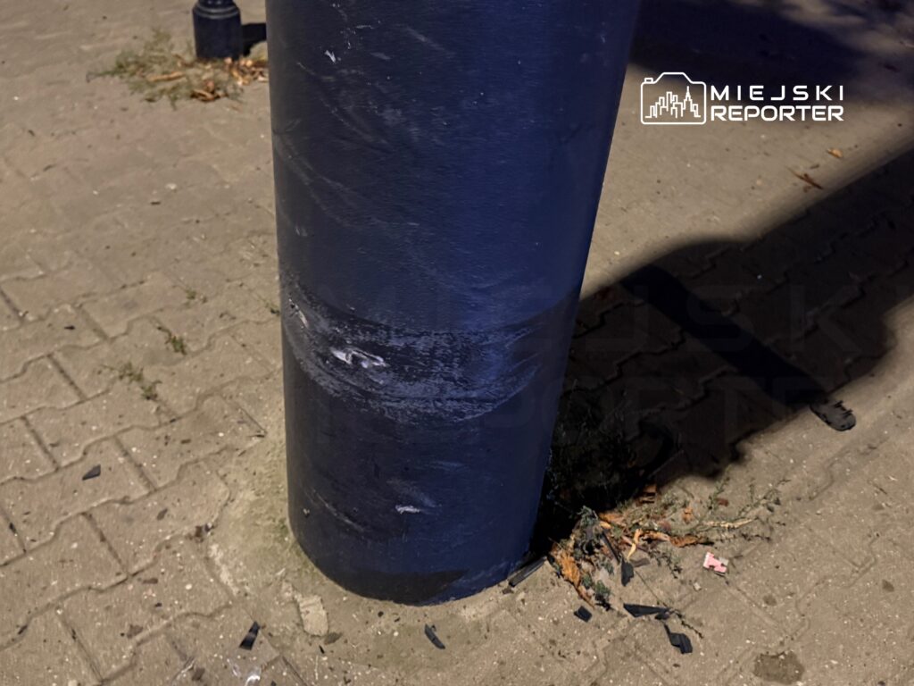 Czarny słup oświetleniowy z widocznymi zadrapaniami, stojący na chodniku w miejskim otoczeniu.