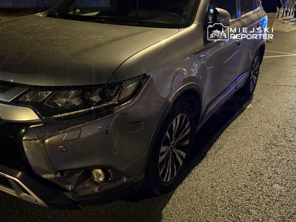 Uszkodzony srebrny SUV zaparkowany na oświetlonej ulicy, z widocznymi zarysowaniami na przedniej części.