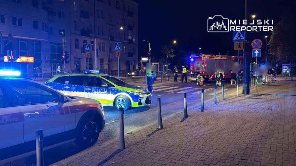Na ulicy nocą, policjanci i strażacy interweniują przy zdarzeniu, obok zaparkowanych radiowozów z włączonymi sygnałami świetlnymi.