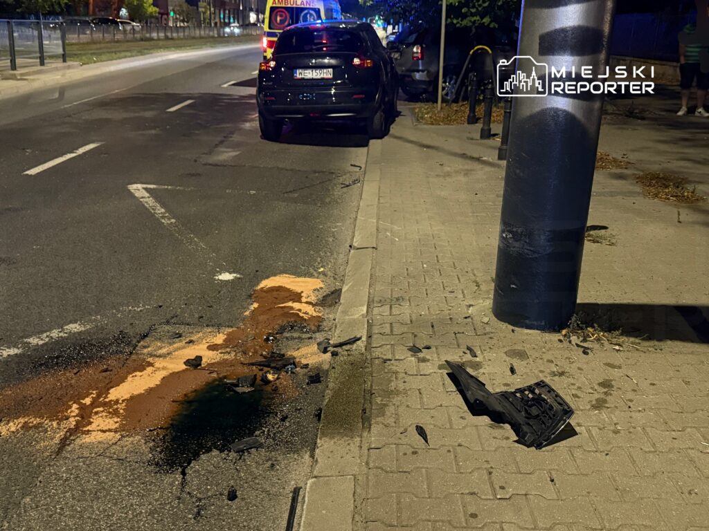 Na jezdni widoczne są ślady płynów oraz uszkodzone fragmenty pojazdu obok słupa. W tle stoi ambulans.