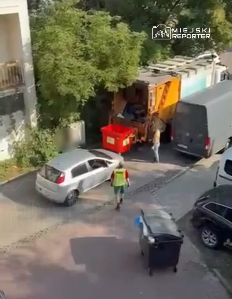 Mężczyzna w kamizelce roboczej zbiera odpady z pojemnika na tle zaparkowanych samochodów.