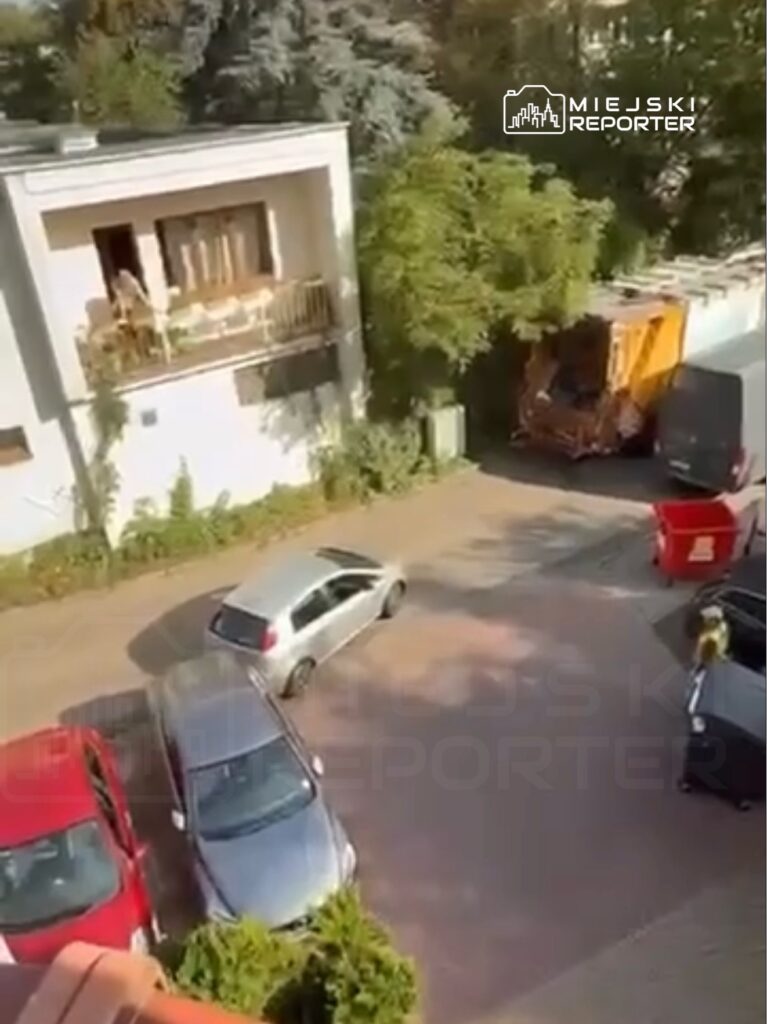 Srebrny samochód osobowy skręca w kierunku parkingu, przy nowoczesnym budynku mieszkalnym z balkonem.