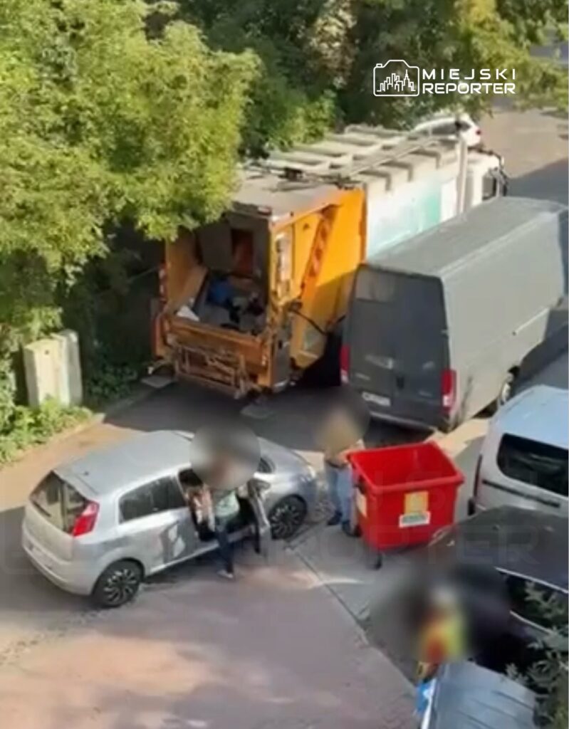 Grupa osób przy samochodzie osobowym rozmawia, podczas gdy śmieciarka opróżnia kontener w tle.