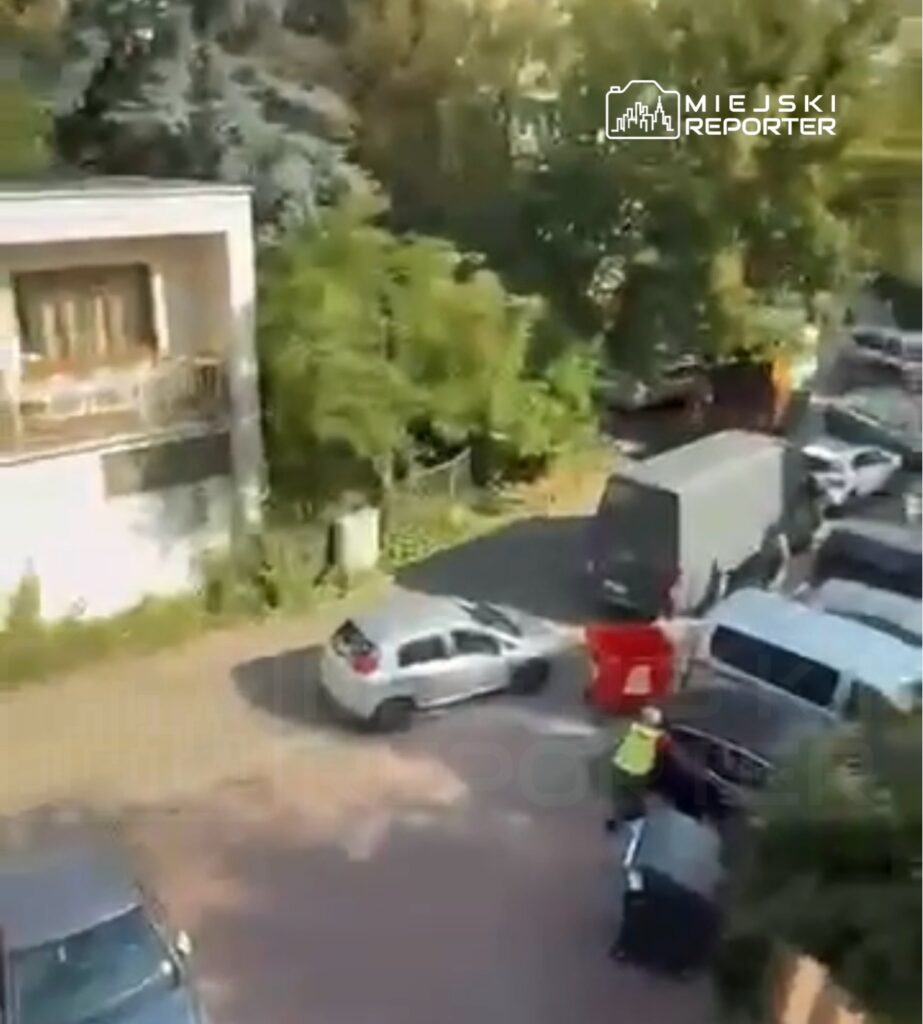 Kierowca samochodu osobowego manewruje na wąskiej ulicy między zaparkowanymi pojazdami.