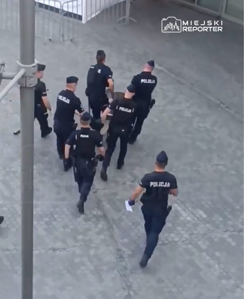 Grupa siedmiu policjantów w mundurach prowadzi osobę na terenie otwartego placu.