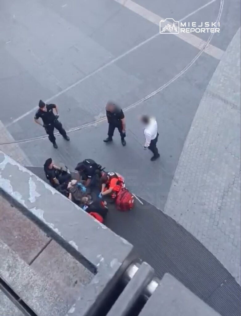 Grupa ratowników i policjantów udziela pomocy osobie leżącej na chodniku w miejskim otoczeniu.