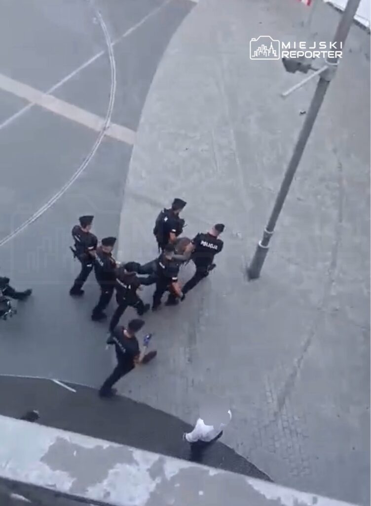 Grupa policjantów w mundurach zatrzymuje mężczyznę na miejskim placu, obok znajduje się przechodzień w białej koszuli.